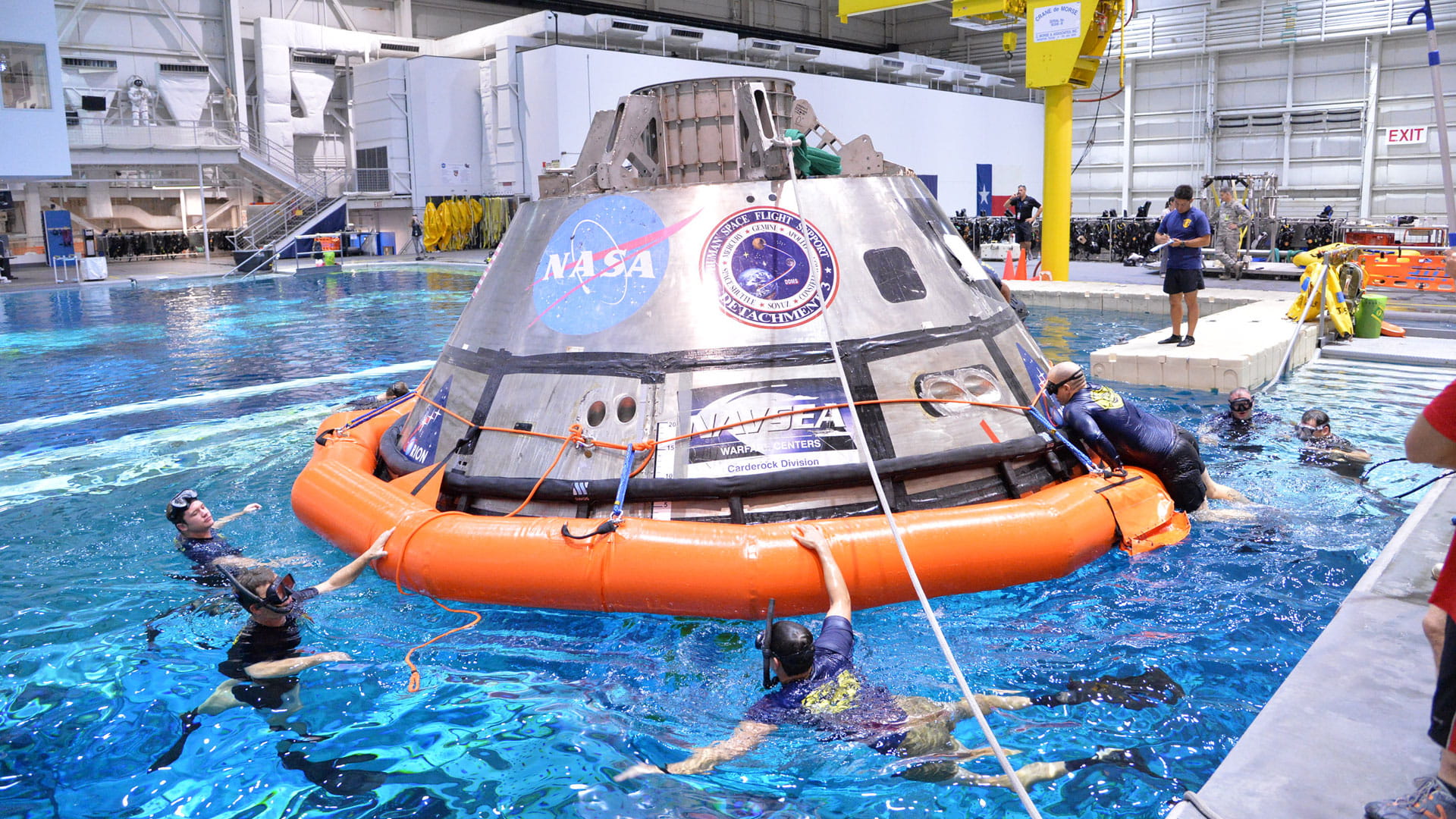 Men in scuba gear surrounding a space capsule in a testing facility's pool