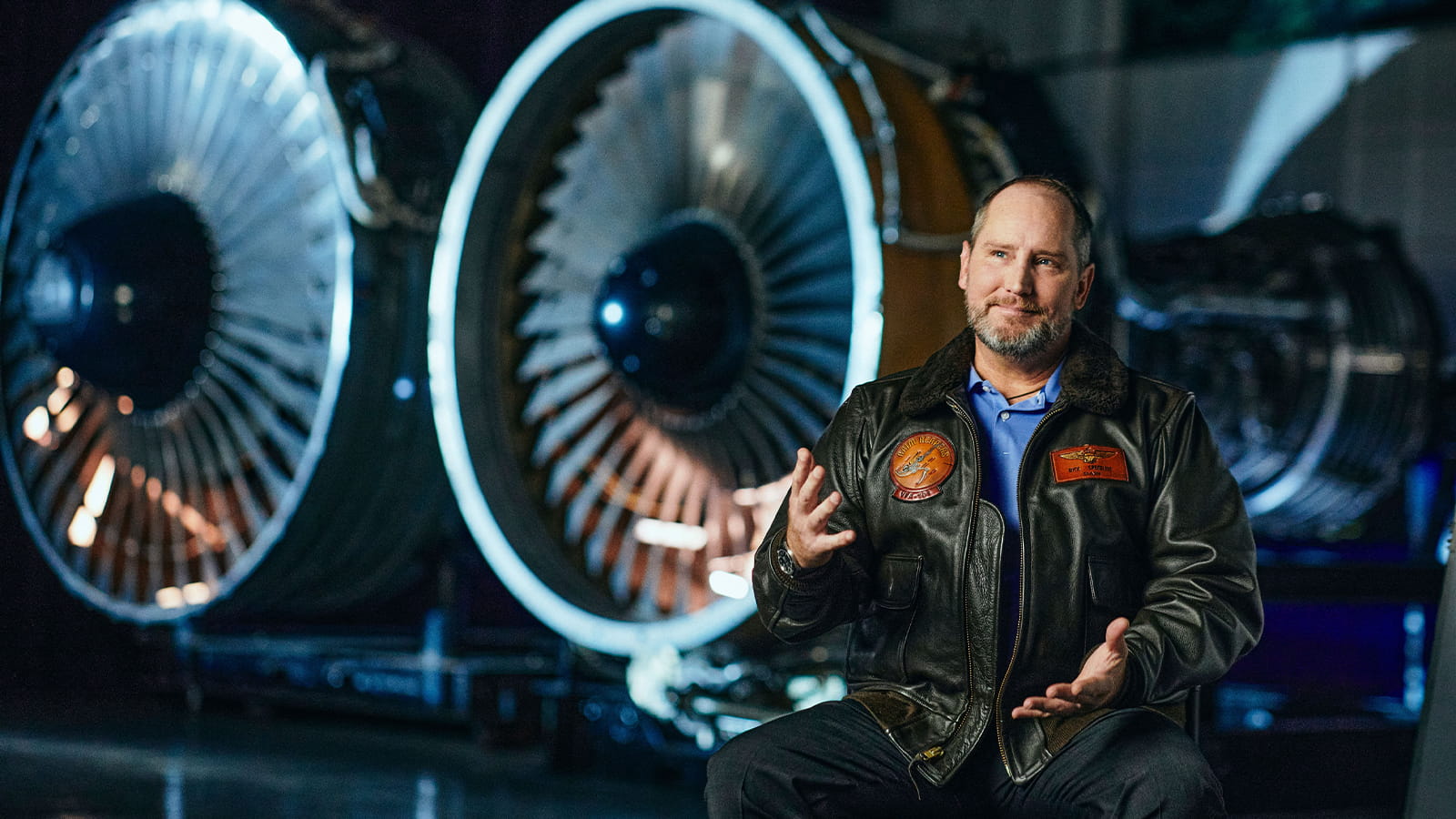 A man in a leather flight jacket gestures in front of two aircraft engines during an interview