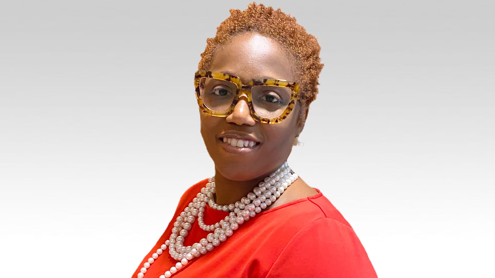 A woman wearing a reddish-orange top, strands of pearls and tortoise shell eyeglasses poses for a professional photograph