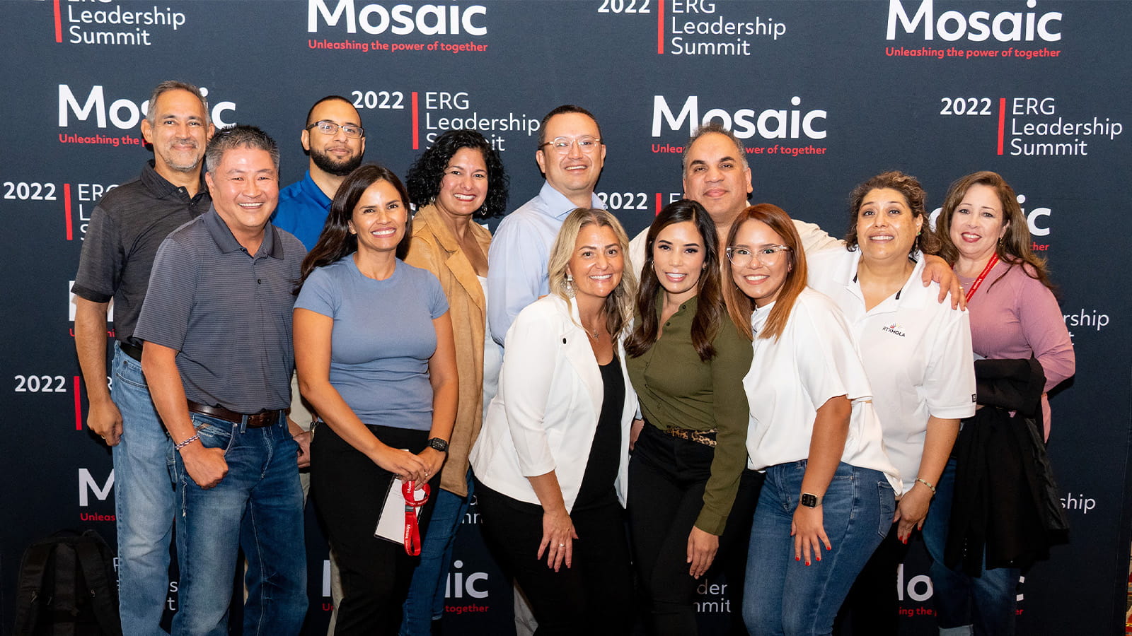 Seven women and five men wearing casual and business-casual clothing smile and pose for a group photo against a dark blue backdrop that reads: Mosaic: Unleashing the power of together, 2022 ERG Leadership Summit