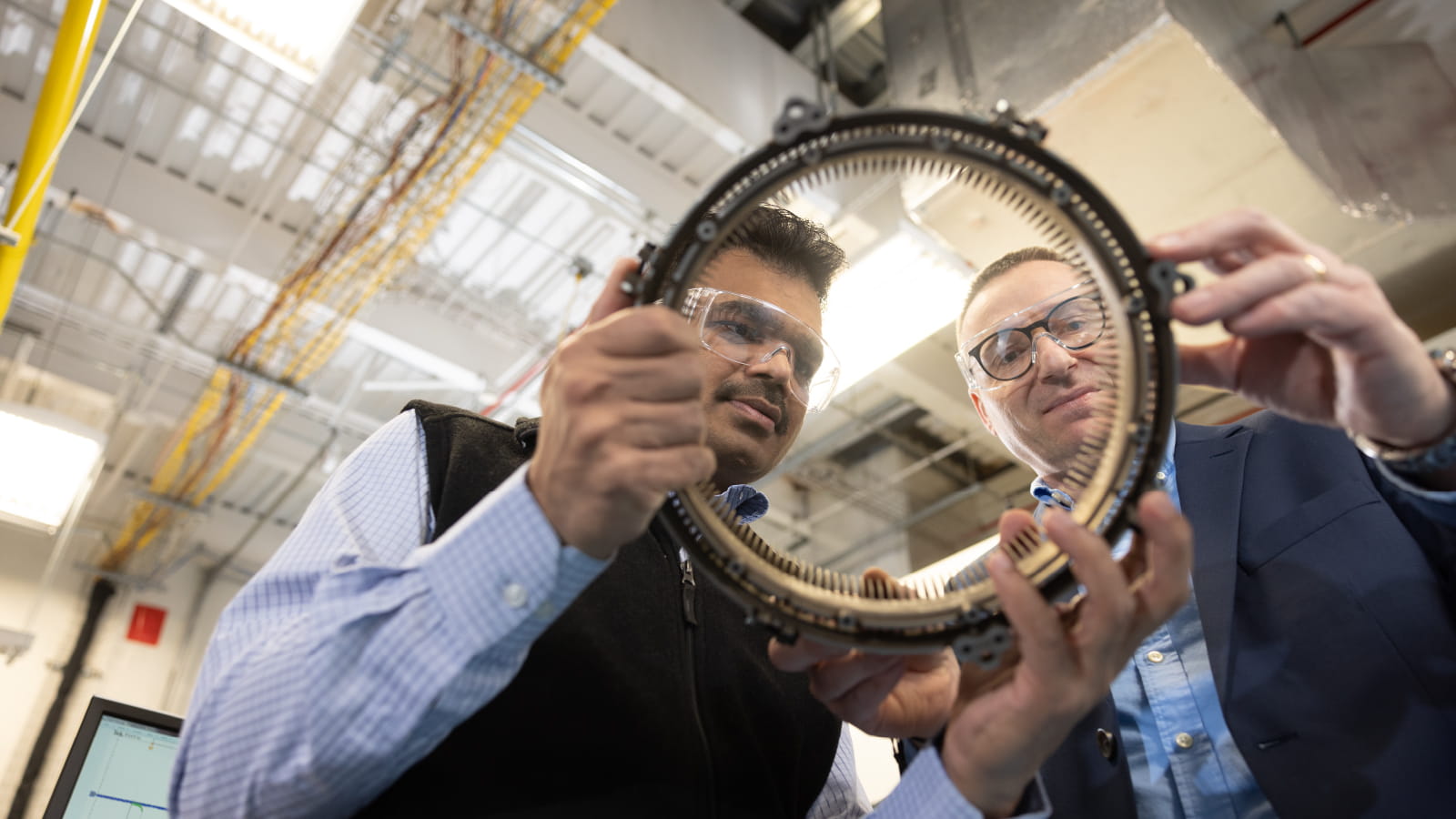 Two male Raytheon Technologies employees wearing protective eye shields holding and inspecting machinery together